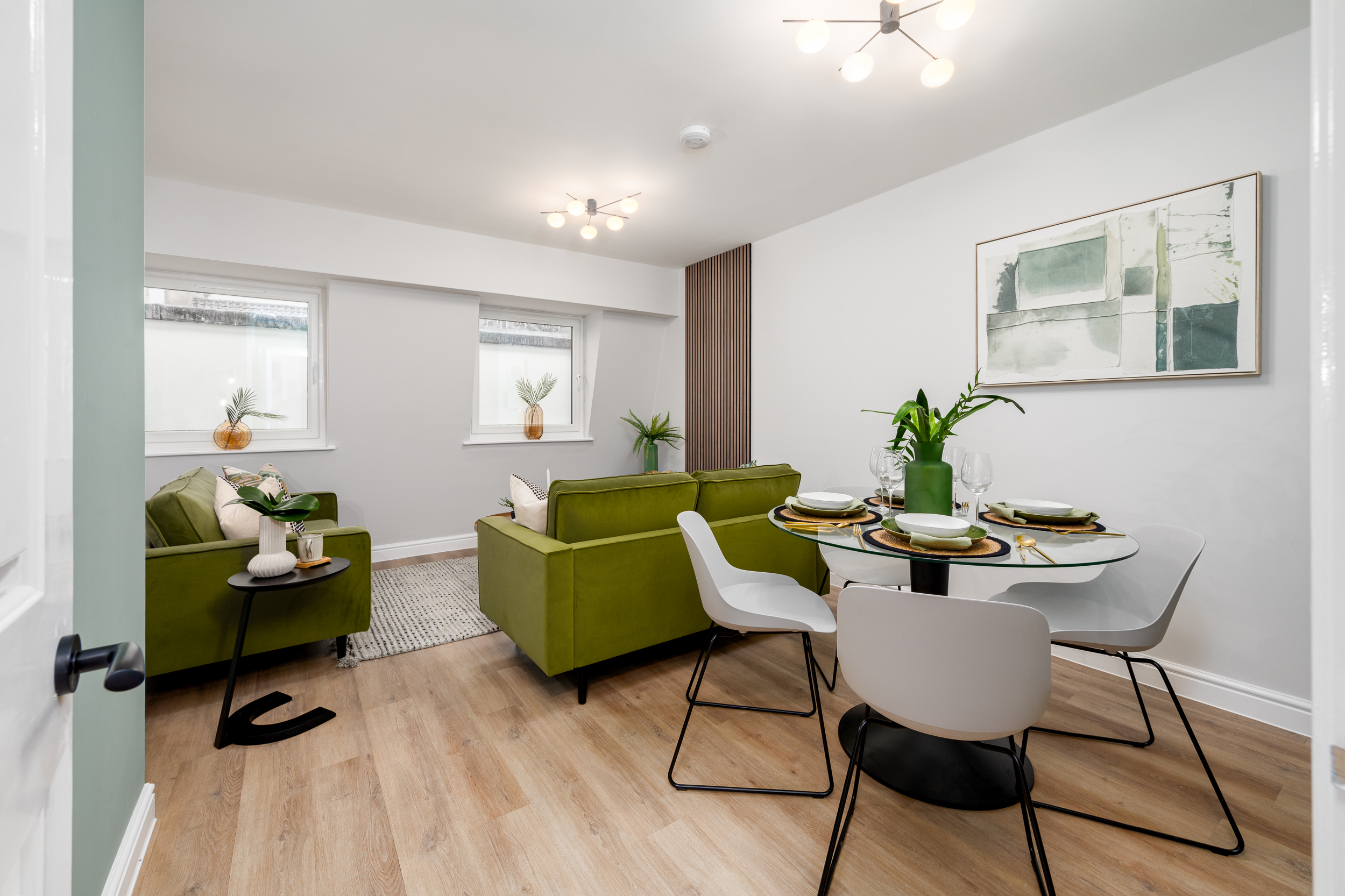 White living area with olive green sofa, glass dining table with grey chairs and feature artwork above.