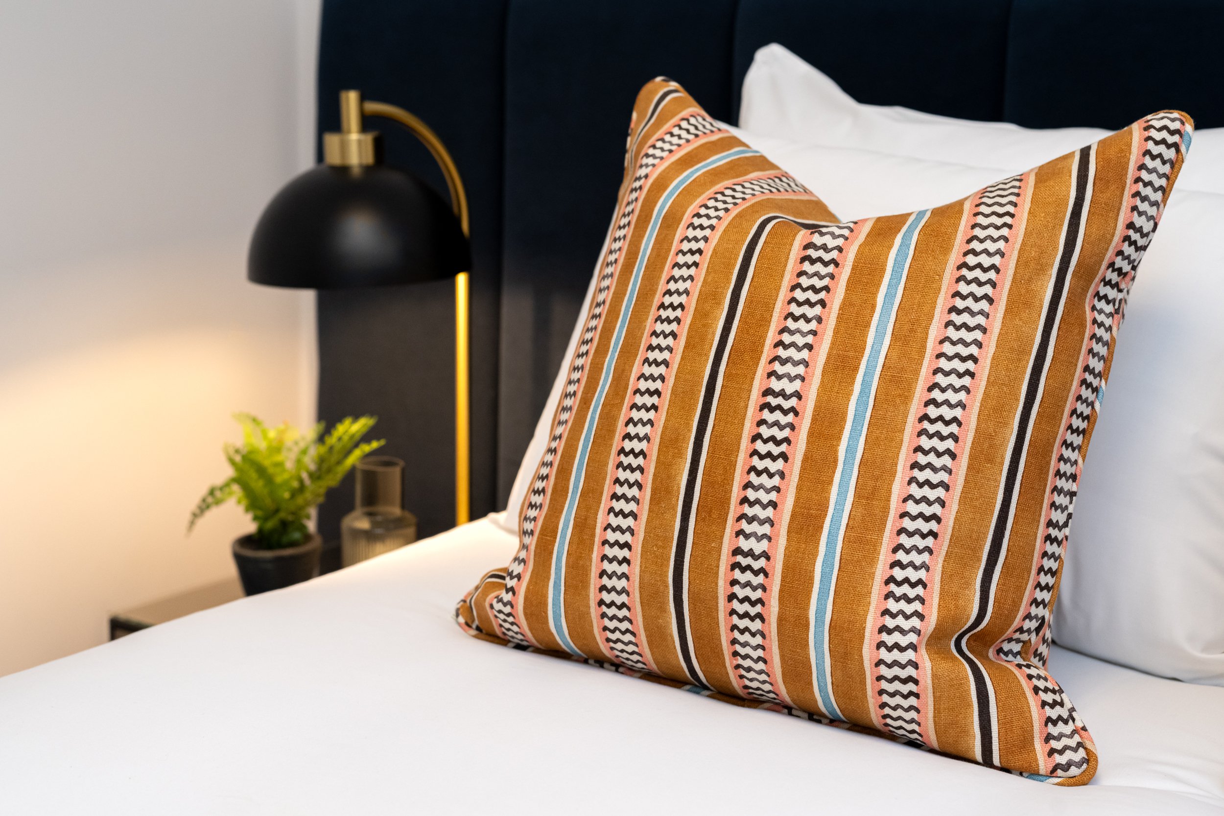 Close up of red and blue cushion on well presented bed with white linen. Black bedside lamp in the background.