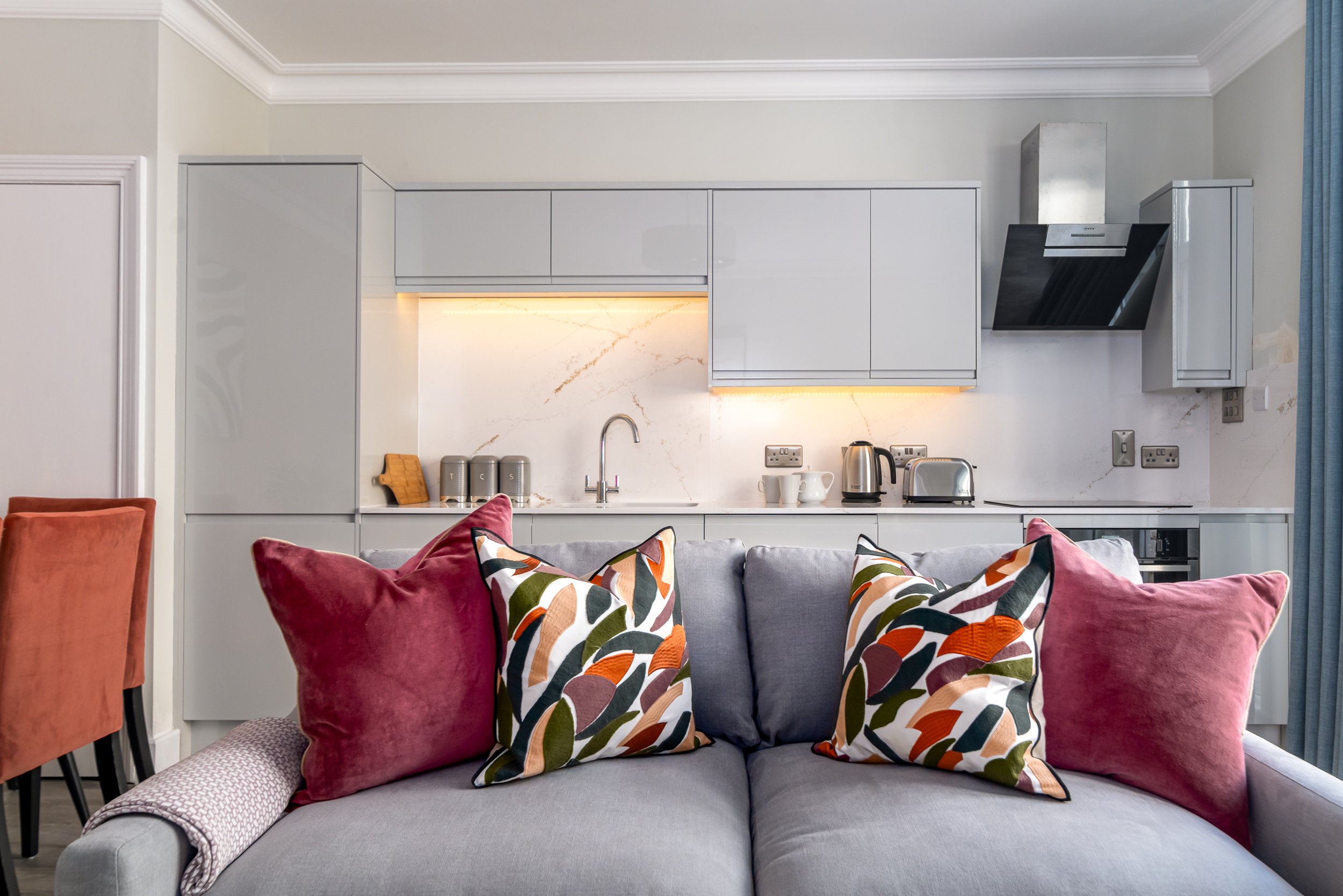Modern kitchen with elegant grey cabinets and a white marble backsplash, accented by colourful throw pillows on a cozy sofa.