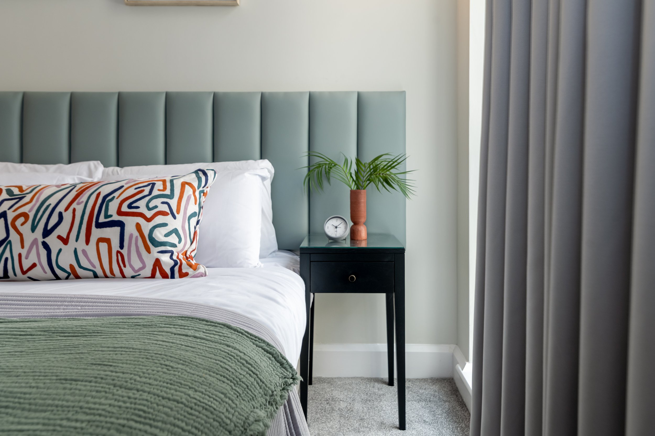 Cosy bedroom featuring a tufted mint green headboard, colourful pillow, bedside table with a clock and plant, and soft grey curtains.
