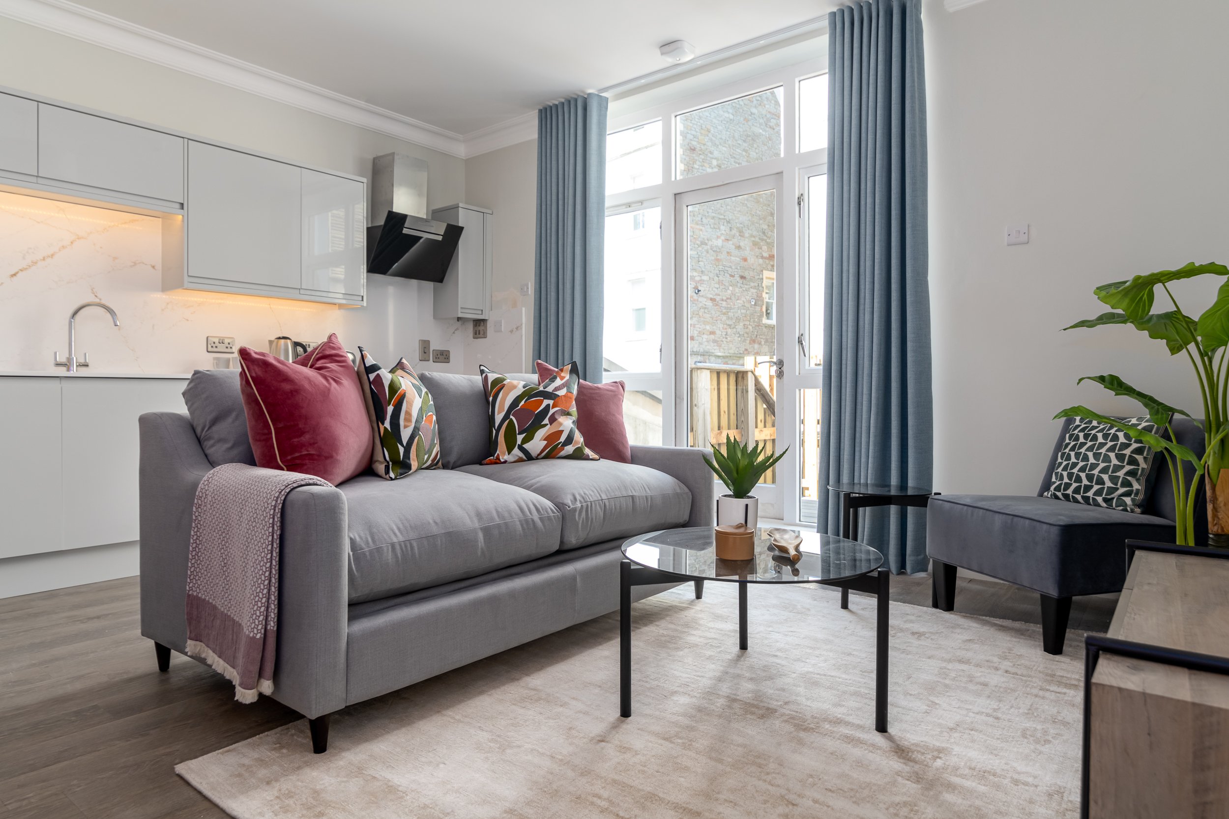 Cosy living room featuring a grey sofa with colourful pillows, a glass coffee table, and large windows with blue curtains.