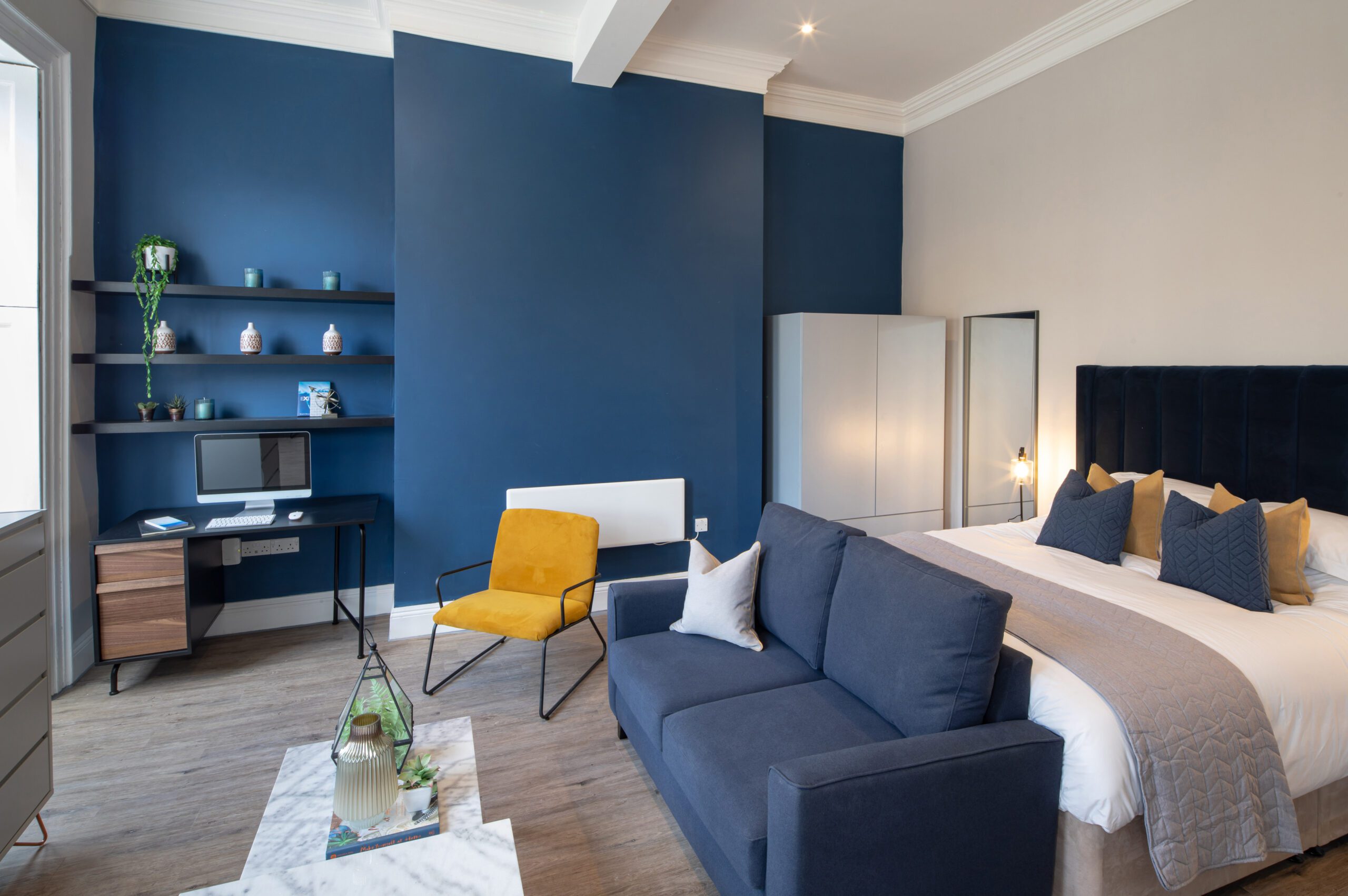 Modern studio apartment with a cozy blue sofa, yellow chair, and a neatly made bed. Blue accent wall features shelves with decor. Bright, inviting space.